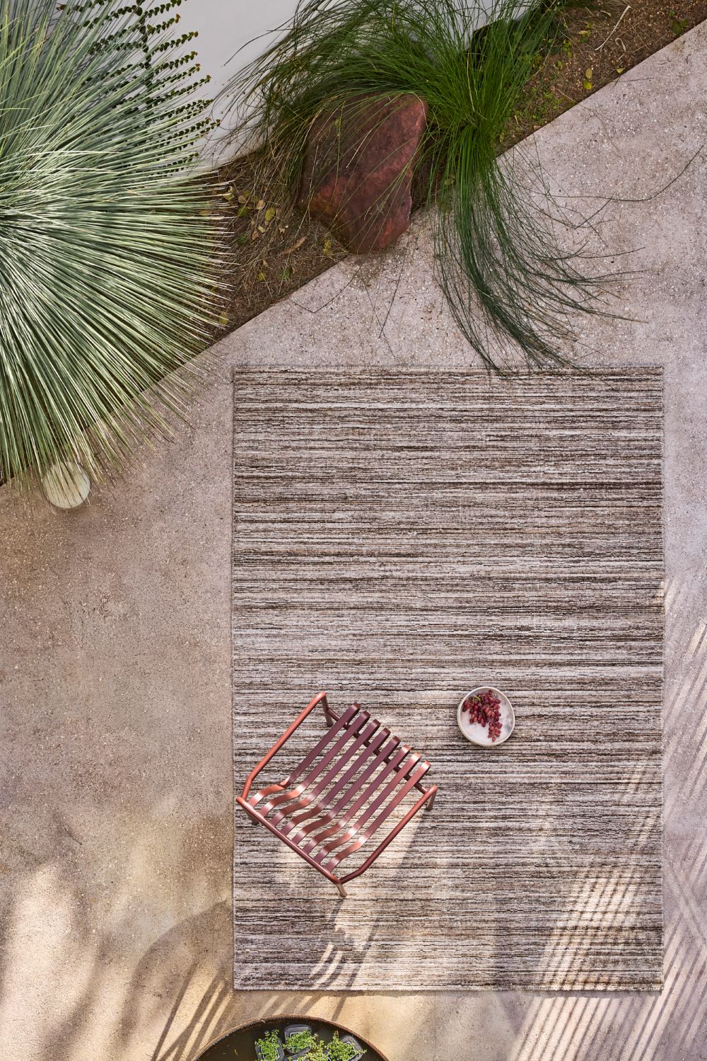 A rectangular outdoor rug with a textured, heathered gray and beige striped pattern, styled on a concrete patio with a small copper wire lounge chair and a decorative bowl.