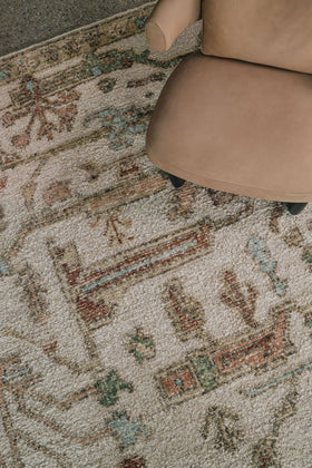 Close-up of the Biraz rug from the Oushak Collection, featuring a traditional pattern in cream, olive, terracotta and turquoise beneath a tan leather chair.