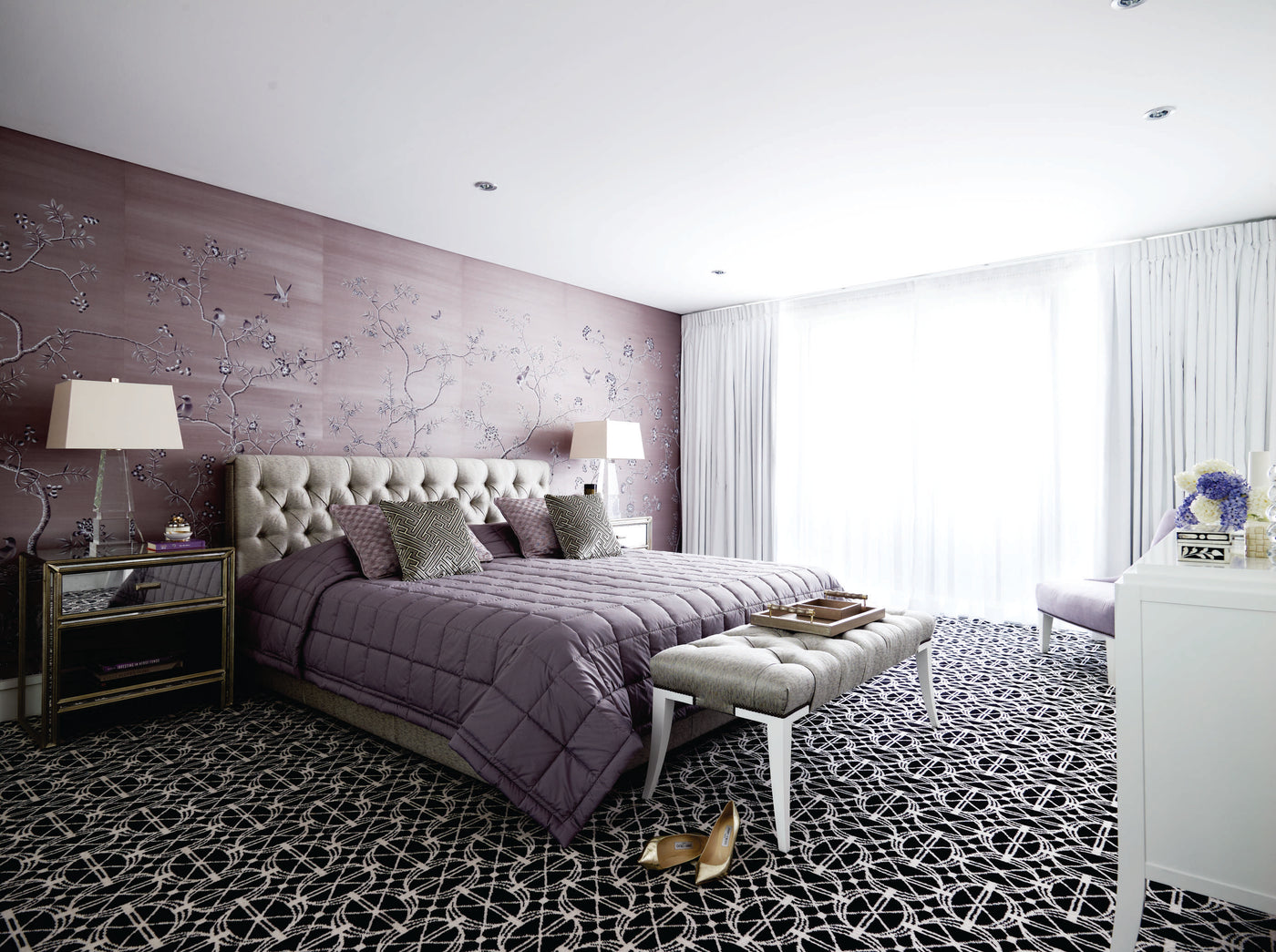 A styled bedroom interior featuring a bed dressed in a quilted mauve-purple bedding set, set against a textured mauve floral accent wall. The room includes a patterned black and white carpet, a tufted bench, bedside tables with lamps, and a white dresser in the foreground.