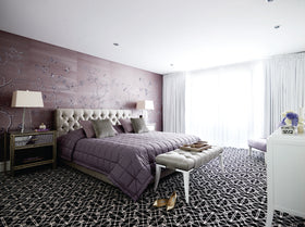 A styled bedroom interior featuring a bed dressed in a quilted mauve-purple bedding set, set against a textured mauve floral accent wall. The room includes a patterned black and white carpet, a tufted bench, bedside tables with lamps, and a white dresser in the foreground.