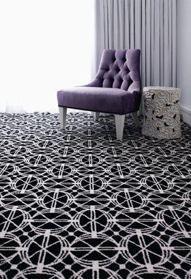 A black and white geometric patterned area rug in a styled room setting, featuring a purple tufted armchair and a carved white side table.