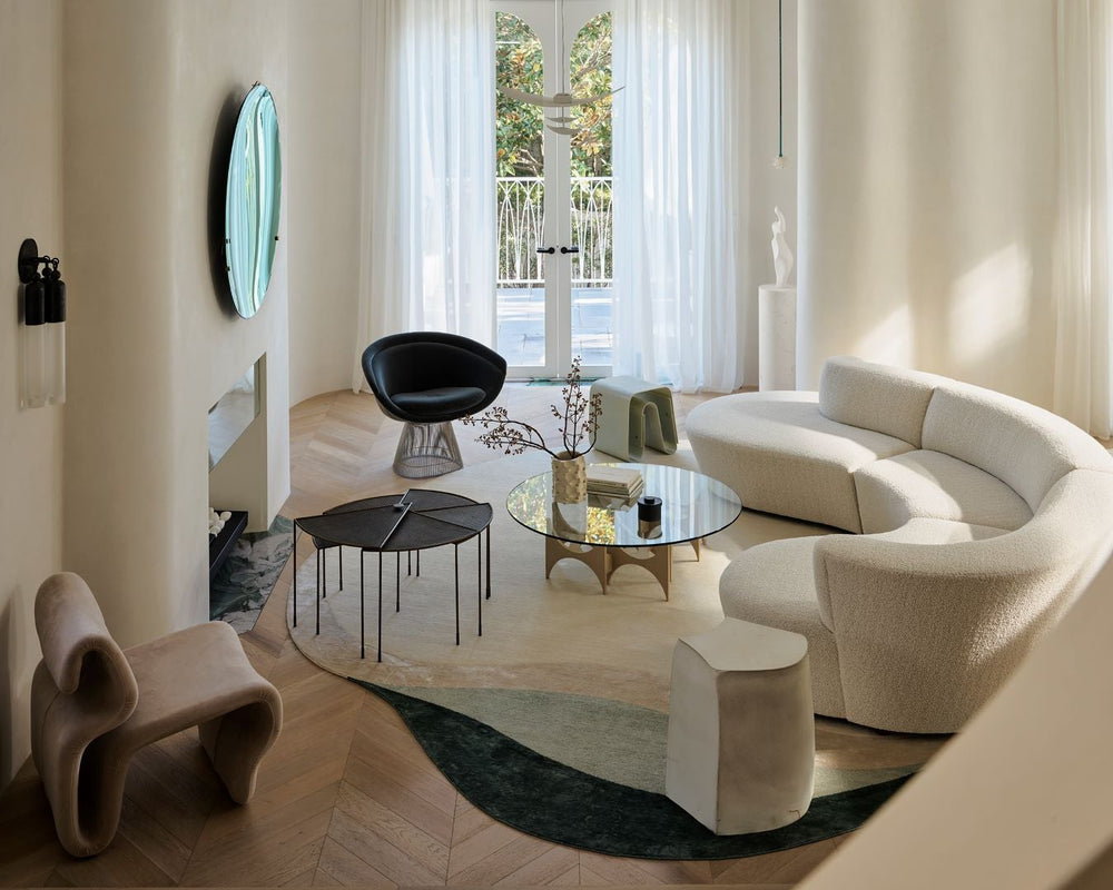 A modern, stylishly designed living room featuring a curved cream bouclé sofa, a sculptural black armchair, a glass-top coffee table, and a dark green abstract-patterned rug on light wood herringbone flooring.