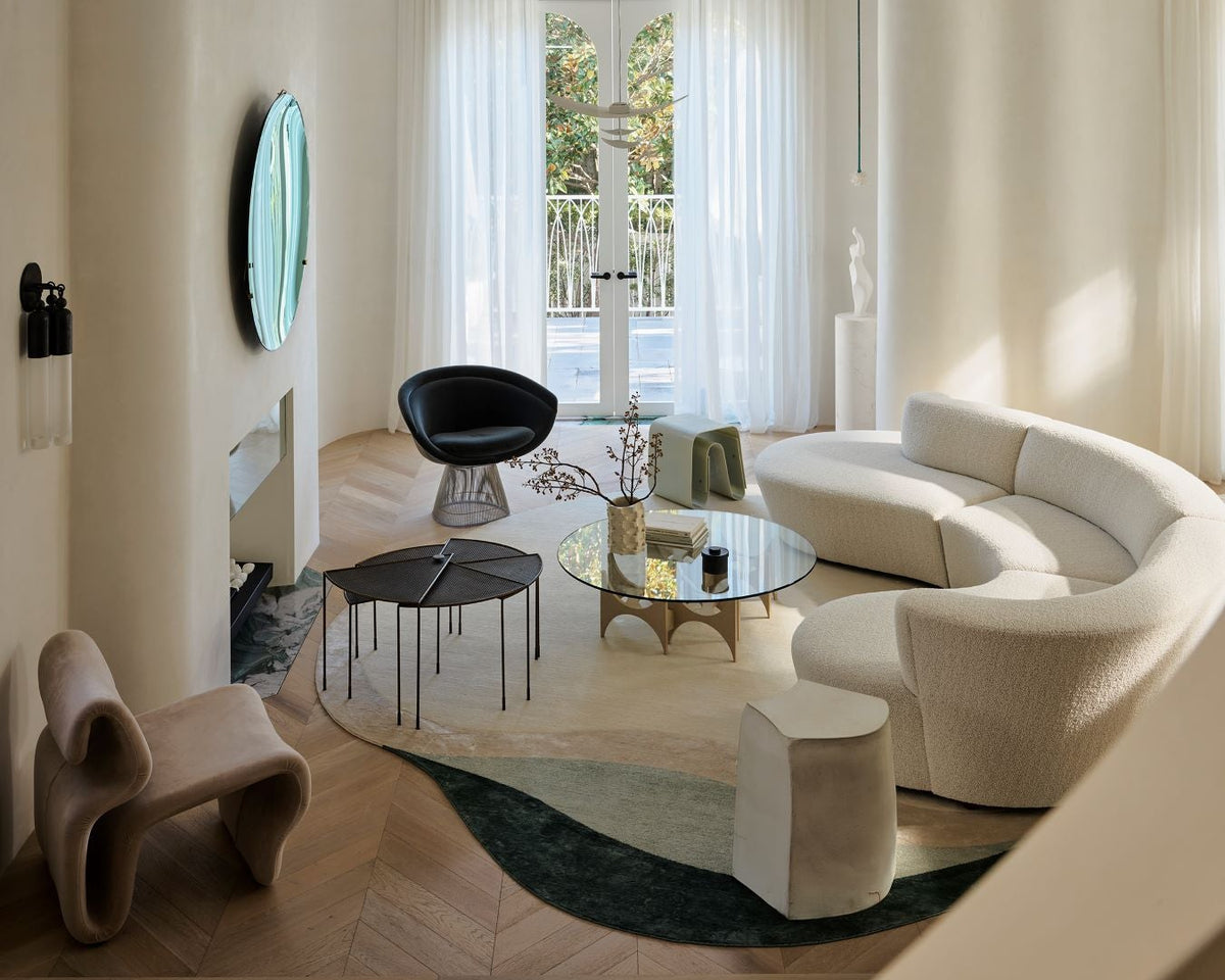 A modern, stylishly designed living room featuring a curved cream bouclé sofa, a sculptural black armchair, a glass-top coffee table, and a dark green abstract-patterned rug on light wood herringbone flooring.