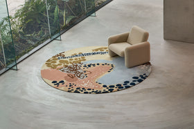 A modern round area rug with an abstract organic pattern in beige, pink, blue, and black, styled in a minimalist interior next to a beige armchair.