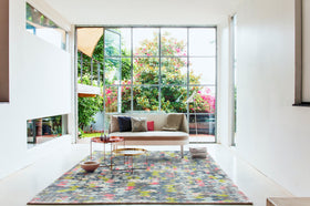 A modern living room with a large floral area rug, a low beige sofa with accent pillows, and two round nesting coffee tables, set in front of a large grid-style glass window overlooking a lush green garden.