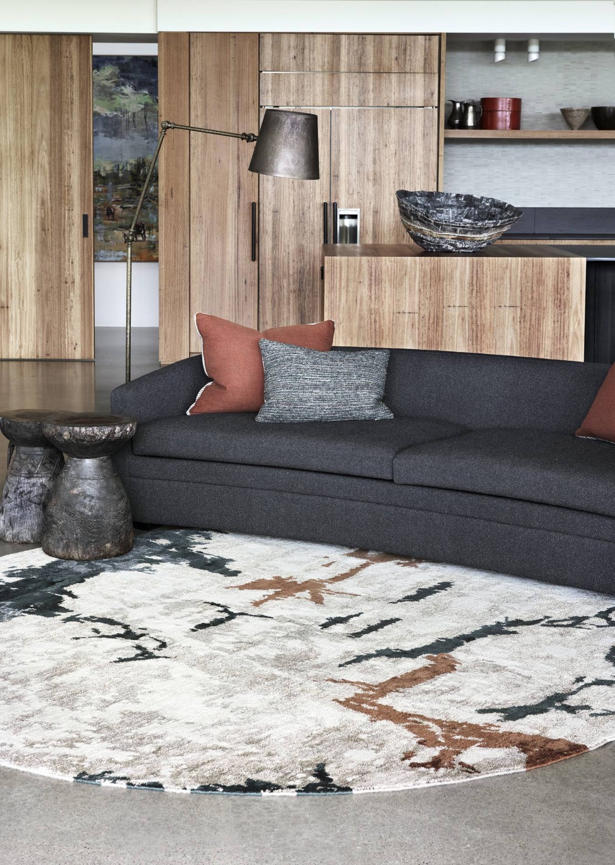 A modern living room scene featuring a dark charcoal gray sectional sofa with rust and gray accent pillows, set on a large round area rug with an abstract black, white, and brown pattern. The background shows light wood cabinetry and a contemporary floor lamp.