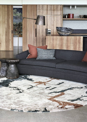 A modern living room scene featuring a dark charcoal gray sectional sofa with rust and gray accent pillows, set on a large round area rug with an abstract black, white, and brown pattern. The background shows light wood cabinetry and a contemporary floor lamp.