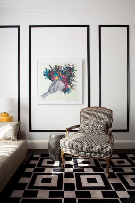 A framed art print featuring a colourful bird illustration is displayed on a white wall in a modern living room setting with a patterned armchair and a black-and-white geometric rug.