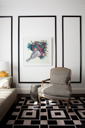 A framed art print featuring a colourful bird illustration is displayed on a white wall in a modern living room setting with a patterned armchair and a black-and-white geometric rug.