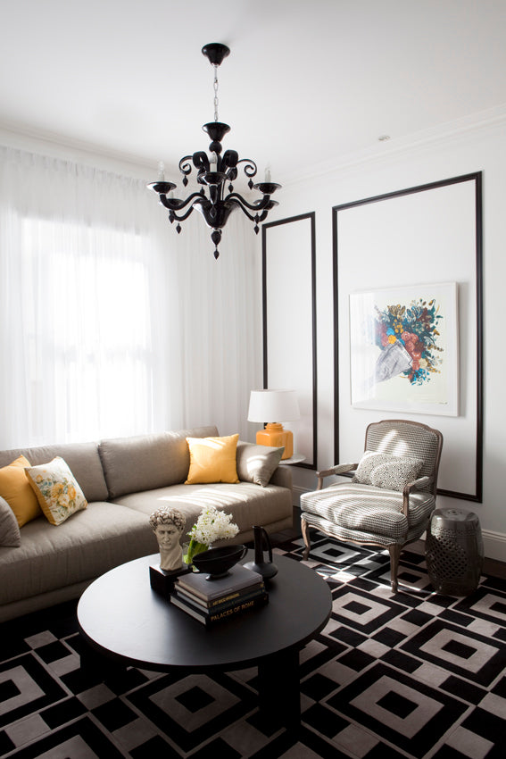 A styled living room featuring a beige sofa with yellow accent pillows, a black round coffee table, a black and white geometric pattern rug, a striped armchair, and a black chandelier.