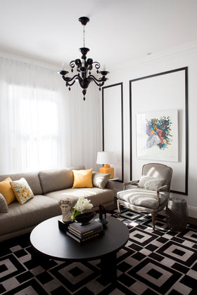 A styled living room featuring a beige sofa with yellow accent pillows, a black round coffee table, a black and white geometric pattern rug, a striped armchair, and a black chandelier.