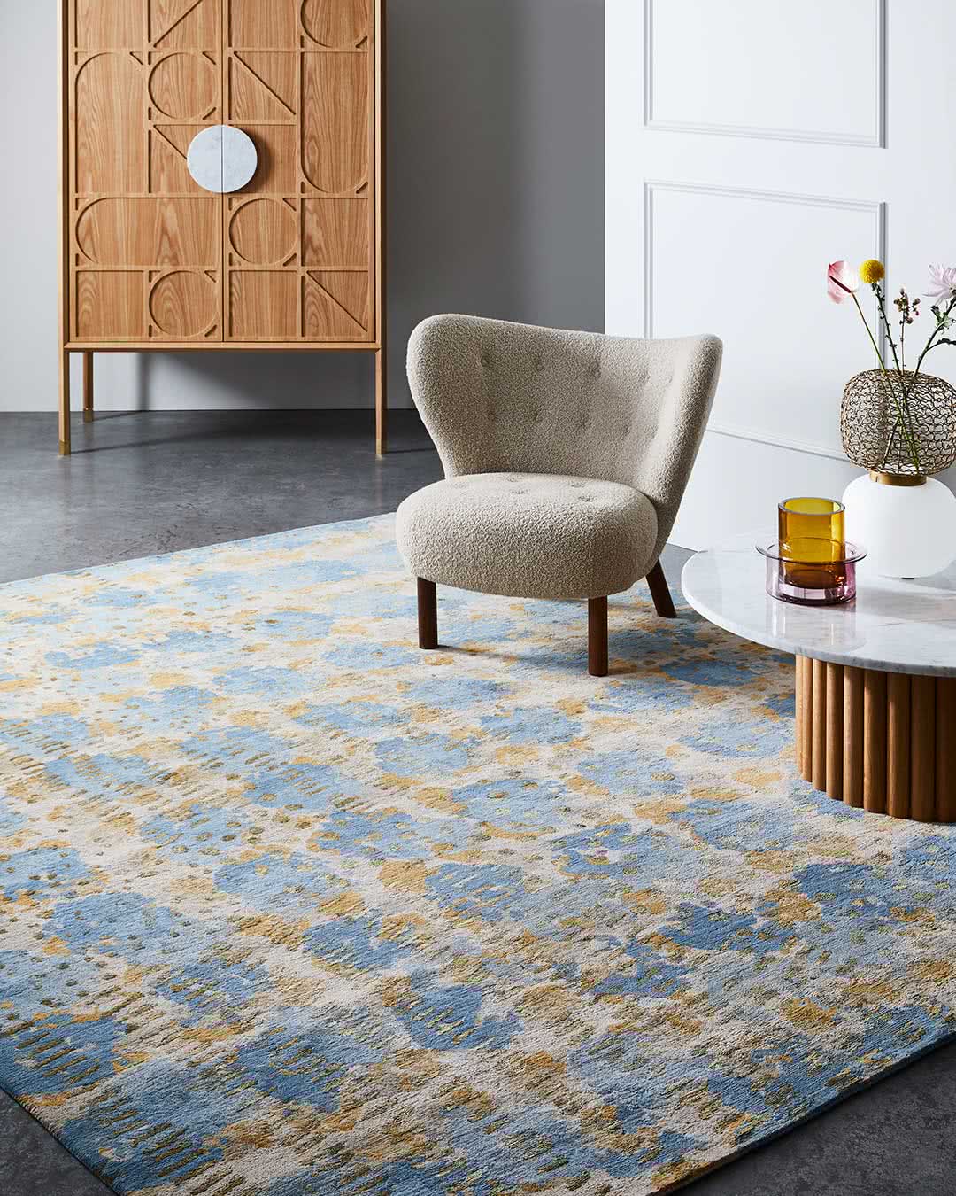 A large area rug with an abstract distressed pattern in blue, beige, and gold tones, styled in a modern living room setting next to a tufted armchair and a marble-top side table.