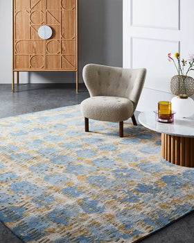 A large area rug with an abstract distressed pattern in blue, beige, and gold tones, styled in a modern living room setting next to a tufted armchair and a marble-top side table.