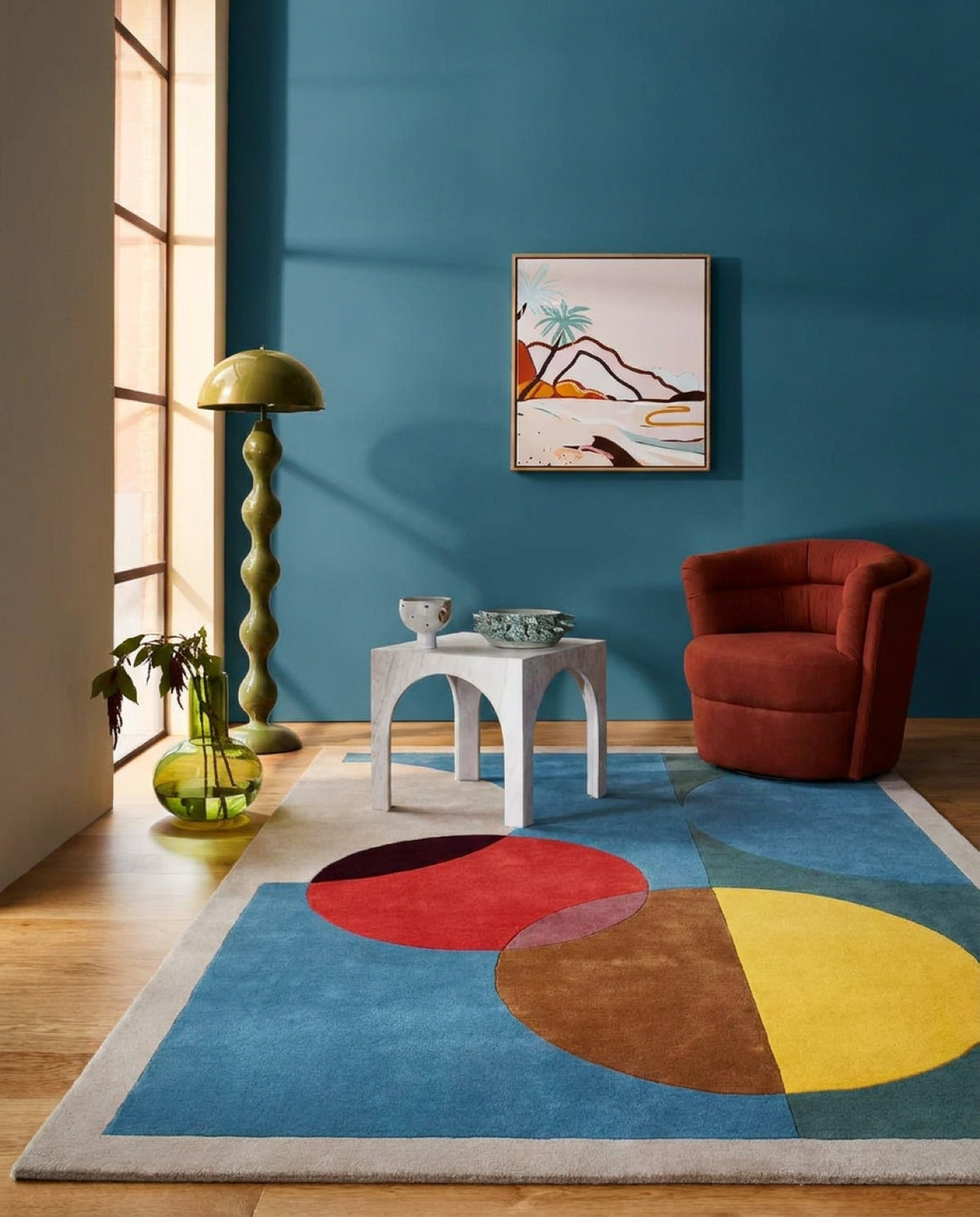A modern living room scene featuring a prominent area rug with a bold geometric color-block pattern in blue, red, yellow, brown, and beige. The room is styled with a terracotta armchair, a white side table, a green spiral floor lamp, and a blue wall with abstract wall art.