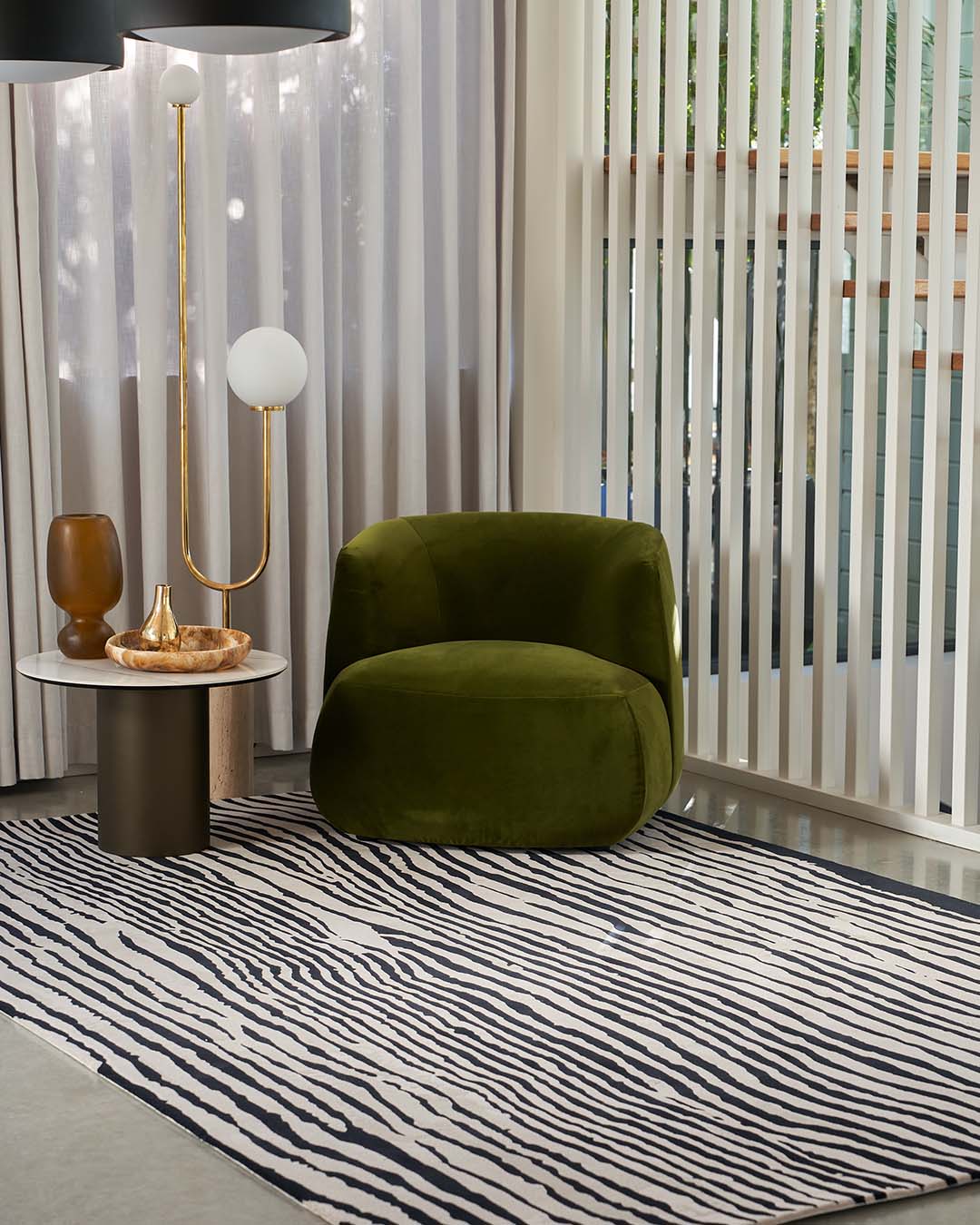 An olive green velvet armchair in a modern living room setting with a black and white zebra-print rug, a gold floor lamp, and vertical white slat screens.