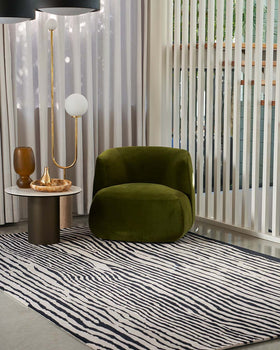 An olive green velvet armchair in a modern living room setting with a black and white zebra-print rug, a gold floor lamp, and vertical white slat screens.