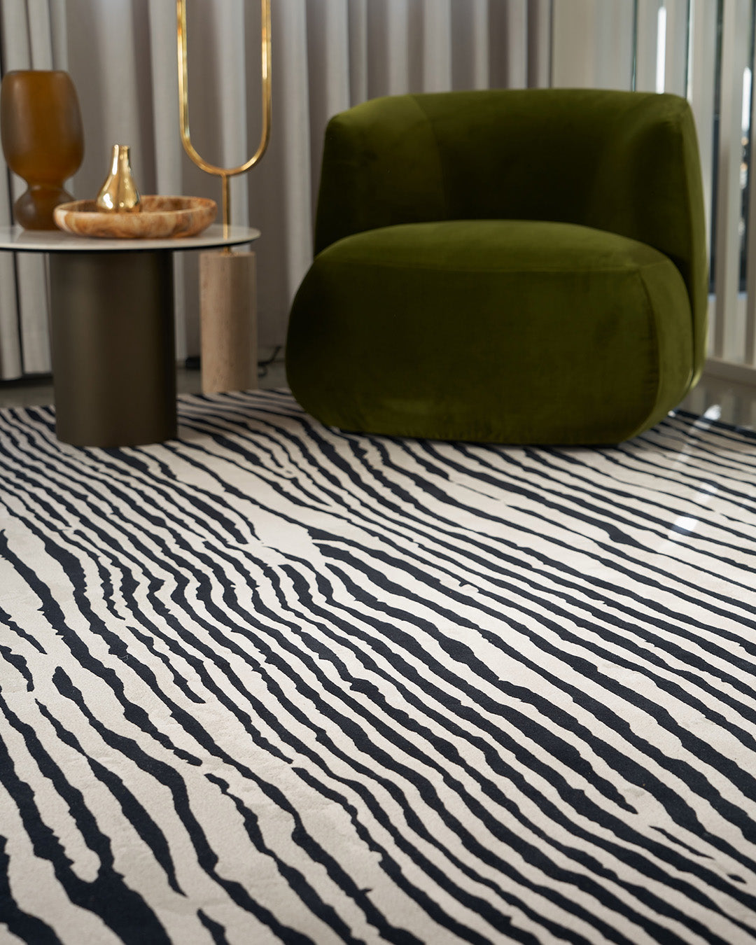 A modern living room scene featuring a prominent black and white zebra-print area rug, with a plush olive green armchair and a small side table in the background.