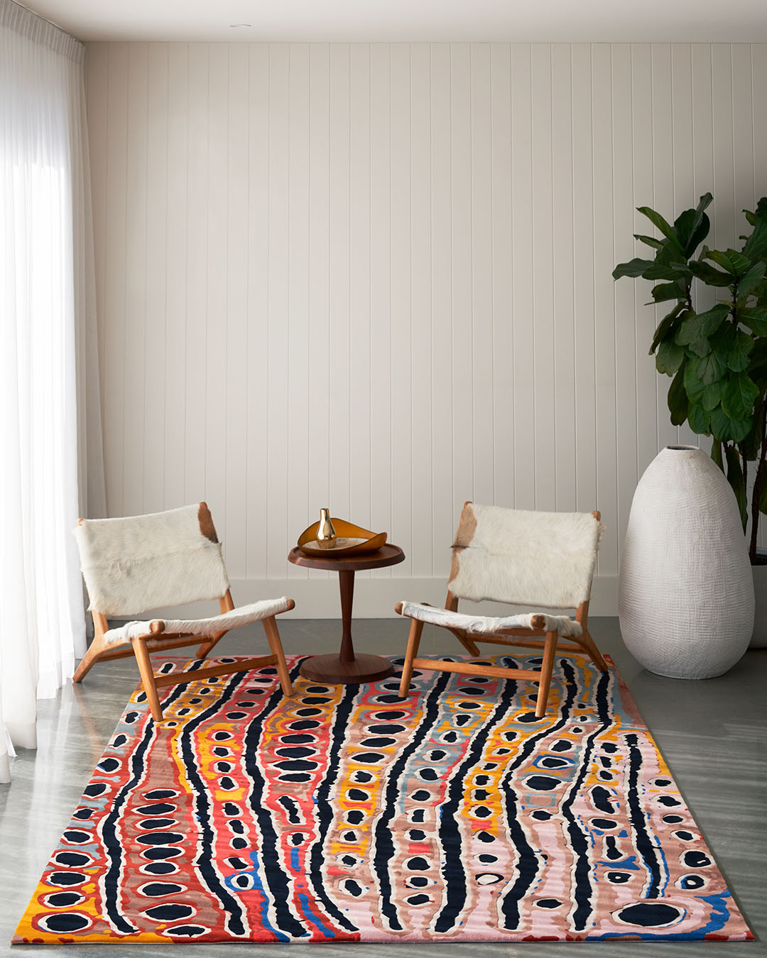 A colorful area rug with abstract wavy stripes in orange, pink, black, blue, and yellow, styled in a modern living room setting with two mid-century modern chairs, a small wooden table, and a large potted plant.