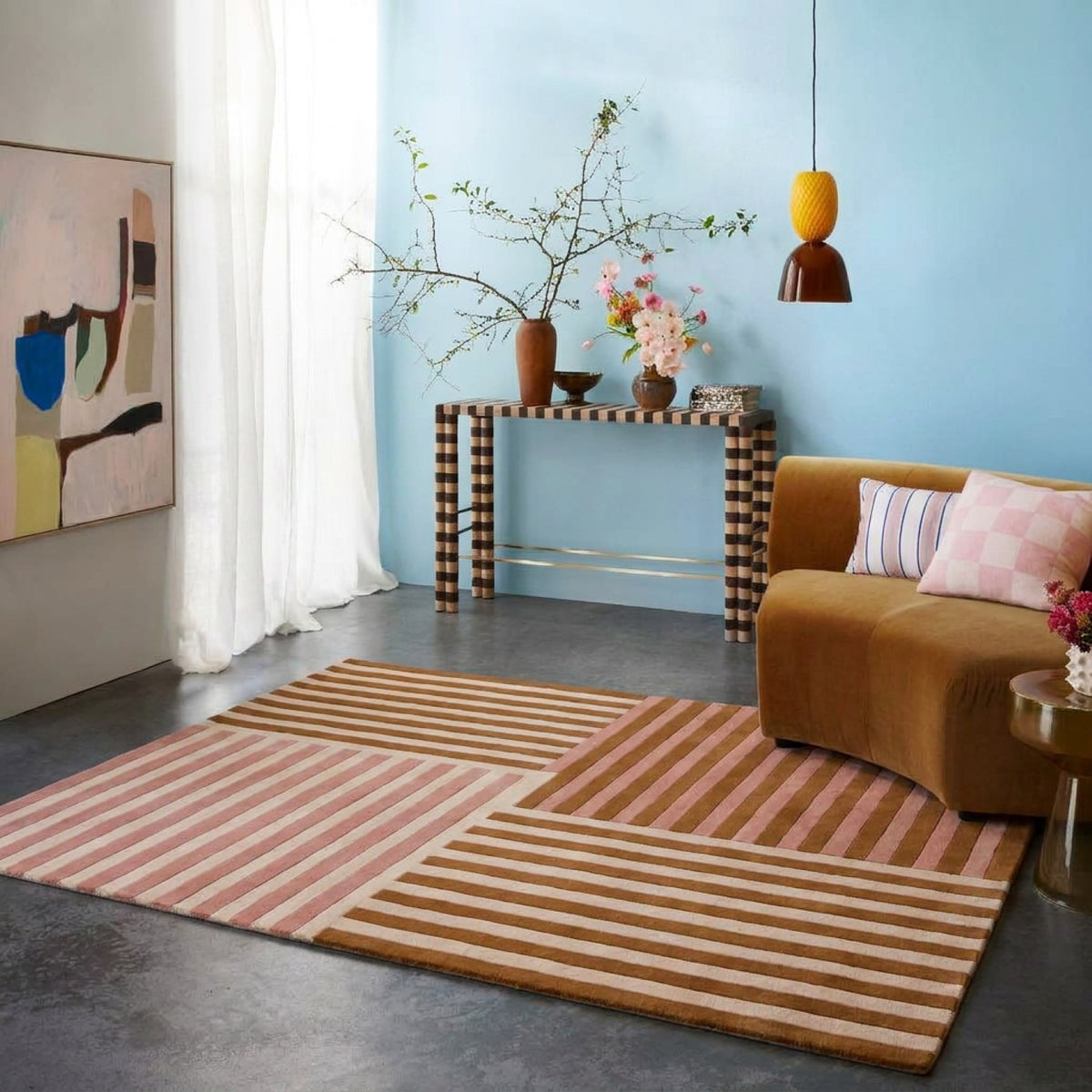 A styled living room scene featuring a large rectangular area rug with a geometric patchwork design of pink, beige, and brown stripes. The room has a light blue wall, a brown velvet sofa with pink and white pillows, a black and white checkered console table, and a modern abstract painting.