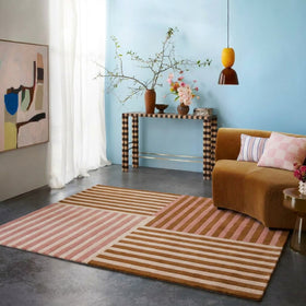 A styled living room scene featuring a large rectangular area rug with a geometric patchwork design of pink, beige, and brown stripes. The room has a light blue wall, a brown velvet sofa with pink and white pillows, a black and white checkered console table, and a modern abstract painting.