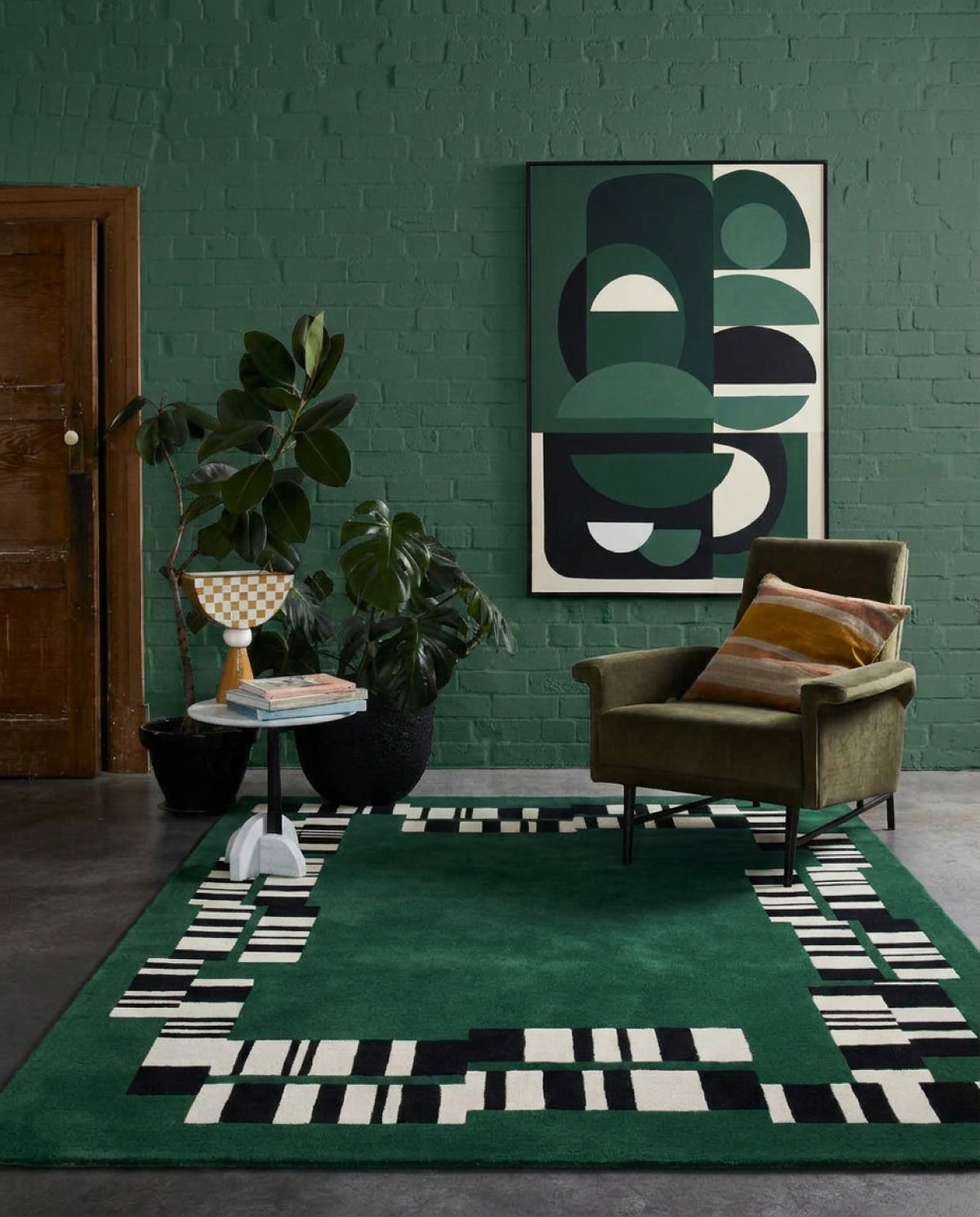 A green area rug with a black and white geometric border pattern is styled in a modern living room setting with a green brick wall, an olive green armchair, and a large abstract art print.