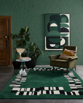 A green area rug with a black and white geometric border pattern is styled in a modern living room setting with a green brick wall, an olive green armchair, and a large abstract art print.