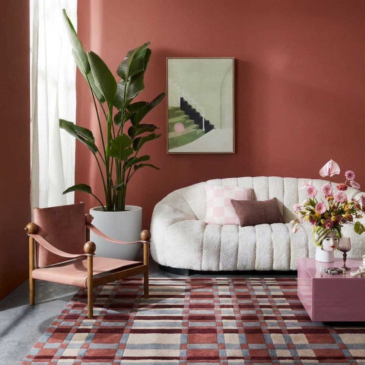 A styled living room corner with a terracotta-pink wall, a curved white boucle sofa with pink and brown pillows, a pink accent chair, a large potted plant, a plaid rug, and a pink side table with flowers.