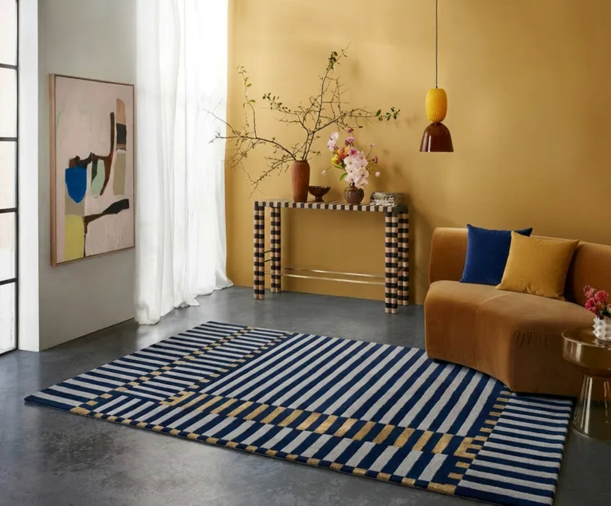 A modern area rug with navy blue, white, and mustard yellow stripes and geometric border details, styled in a contemporary living room with a mustard yellow wall, brown sofa, and decorative console table.