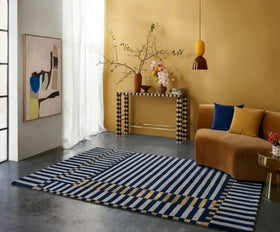 A modern area rug with navy blue, white, and mustard yellow stripes and geometric border details, styled in a contemporary living room with a mustard yellow wall, brown sofa, and decorative console table.