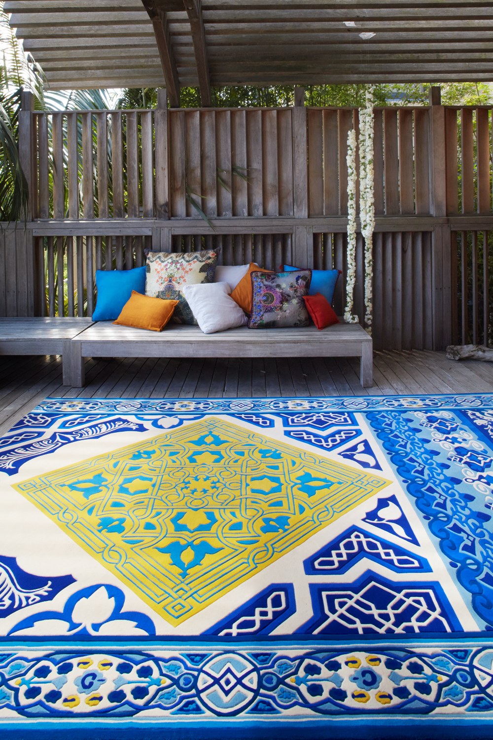 A large Moroccan-style area rug with a blue and yellow geometric pattern is displayed on a wooden deck in front of a wooden bench with colorful throw pillows.