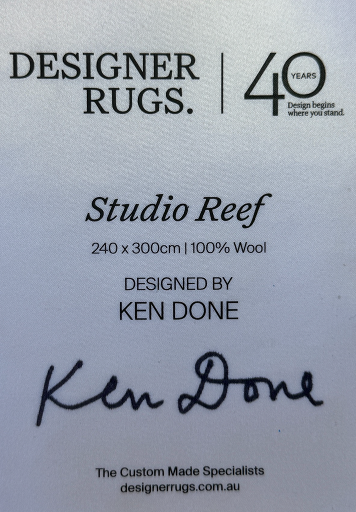 A white product information card for Designer Rugs Studio Reef, showing dimensions, wool content, designer Ken Done, and a signature, on a white background.