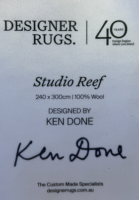 A white product information card for Designer Rugs Studio Reef, showing dimensions, wool content, designer Ken Done, and a signature, on a white background.