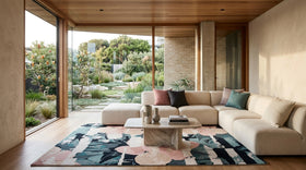 A modern living room with a large beige sectional sofa, a colorful floral rug, and a marble coffee table, set in a minimalist home with large windows overlooking a lush garden.