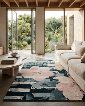 A modern living room featuring a rectangular area rug with a large-scale floral pattern in dusty pink, slate blue, and charcoal gray. The rug is styled on a concrete floor next to a light beige sofa, with large glass doors in the background showing a garden view.